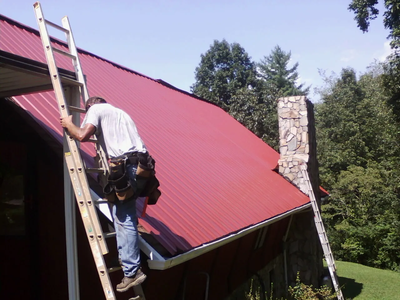 Roofer on a ladder beside a red metal roof and stone chimney — Soffit & Fascia Repair by Roofing Brothers in Blairs